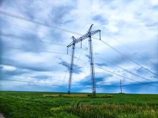 Towering power lines stretch across a vast green field under a dramatic sky with clouds