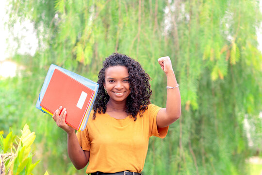 "Estudante brasileira negra feliz comemorando sua aceita&ccedil;&atilde;o na faculdade ao ar livre, segurando cadernos e levantando o bra&ccedil;o com alegria. Conceito de sucesso, motiva&ccedil;&atilde;o e o sonho do ensino superior.