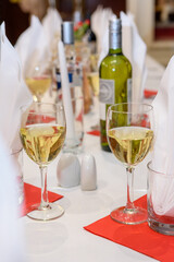 Eleganckie nakrycie stołu z kieliszkami białego wina na czerwonych serwetkach, butelka wina oraz dekoracje w tle. Zdjęcie przedstawia detale stołowej aranżacji idealnej na przyjęcia, kolacje czy elega