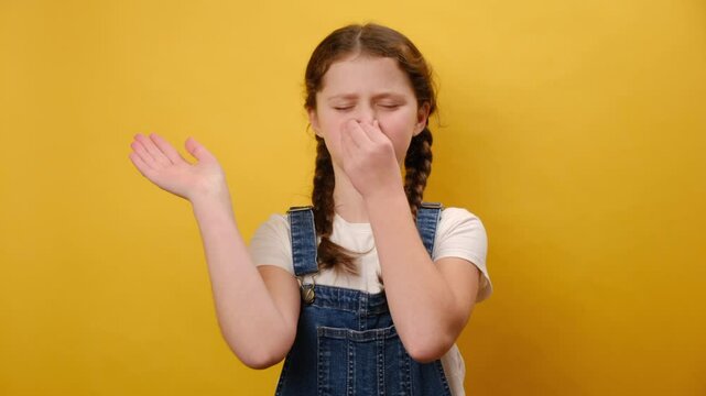 Portrait of preteen girl child holding breath, pinching her nose and expressing disgust to stink, intolerable odor, posing isolated over plain yellow background wall in studio. Childhood concept