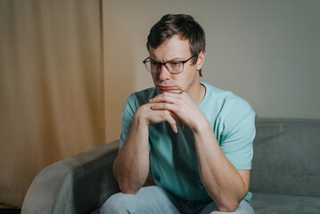 Young man wearing glasses is sitting thoughtfully on a sofa, his hands clasped under his chin, lost...