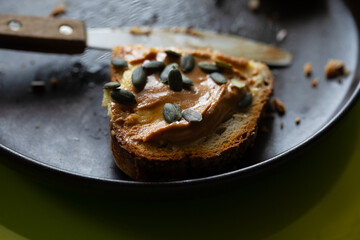 TOASTED BREAD SLICE TOPPED WITH NUT BUTTER AND SCATTERED PUMPKIN SEEDS ON BNROWN STONEWARE PLATE WOODEN HANDLED KNIFE IN BACKGROUND 