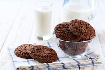 Glass and a jug of fresh milk with chocolate brownie cookies with cracks on a white table, selective focus.