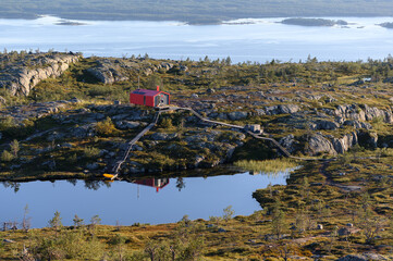 Red cabin on rocky terrain near a calm, reflective pond, surrounded by lush greenery, with a vast lake and distant islands in the background. Peaceful and remote wilderness setting