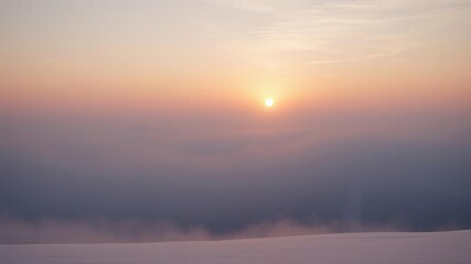 Fototapeta premium The snow-capped mountains are shrouded in a thick layer of fog as the sun rises behind them at dawn, stillness, cloudy sky , daybreak