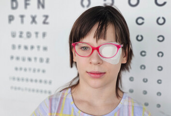 Child with eye patch and glasses in front of eye exam chart at medical clinic concept