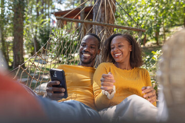 Cheerful African American couple relaxing in a hammock, watching funny videos on a smartphone, enjoying a glamping trip in Europe.