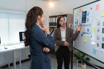 Two asian businesswomen are discussing charts and graphs on a whiteboard during a meeting in a modern office, collaborating on data analysis and strategic planning