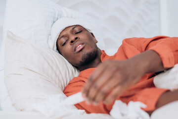 Sick black man lies in bed with a compress on his forehead. Holding a thermometer. Battling fever...