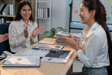 Two happy businesswomen are clapping hands, celebrating successful teamwork and good results in a meeting at the office, surrounded by documents and charts