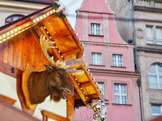 A mounted moose head protrudes from a wooden roof at a festive market, decorated with lights,...