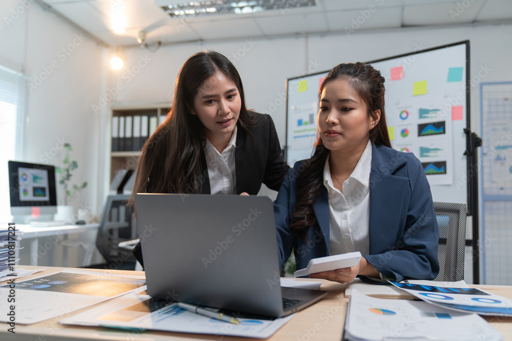 Poster two asian businesswomen are analyzing financial data on a laptop and using a calculator while workin
