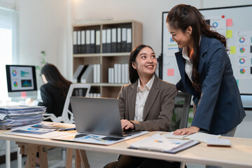 Two smiling asian businesswomen are discussing work while using a laptop in a modern office, collaborating on a project and sharing ideas