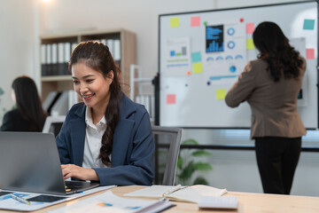Two asian businesswomen are collaborating on a project, with one using a laptop and the other analyzing data on a whiteboard, showcasing teamwork and modern office dynamics