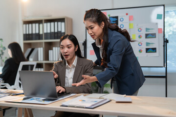 Fototapeta premium Two asian businesswomen are discussing work on a laptop in a modern office, collaborating on a project and analyzing data with surprised expressions