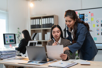 Two asian businesswomen are working together using a laptop and discussing over financial chart documents in a modern office while another colleague is working in the background