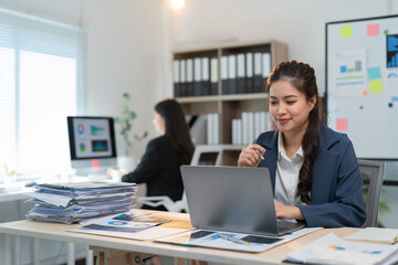 Smiling asian businesswoman analyzing financial charts and typing on a laptop, sitting at her desk in a modern office, her colleague working in the background