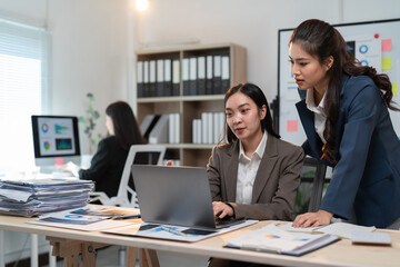 Two asian businesswomen are working together in a modern office, focusing on a laptop and analyzing data while another colleague works in the background