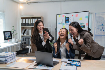Three joyful businesswomen are celebrating a successful project, cheering and laughing around a laptop in a modern office environment, surrounded by charts and paperwork