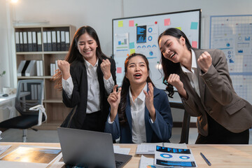Three excited asian businesswomen are celebrating a successful project, raising their fists in joy and cheering in front of a laptop in a modern office meeting