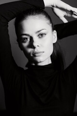 Black and white portrait of a young woman with a serious expression, arms raised, wearing a fitted black turtleneck. Emotional studio setting with soft lighting.