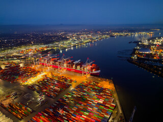 Illuminated Southampton UK container terminal with large container ship at dusk. High altitude aerial view above bright commercial docks with lights towards waterfront city. Industrial backgrounds.