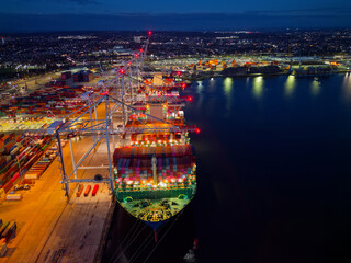 Above bow of large container ship at Southampton Docks UK aerial view. Illuminated container terminal with cranes and lights reflection on calm sea. Beautiful cargo and shipping industrial background.