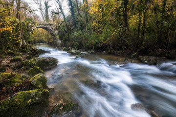 The Alzou river passes under an old stone bridge through a forest of autumn colours near Grammat in the Lot region of France