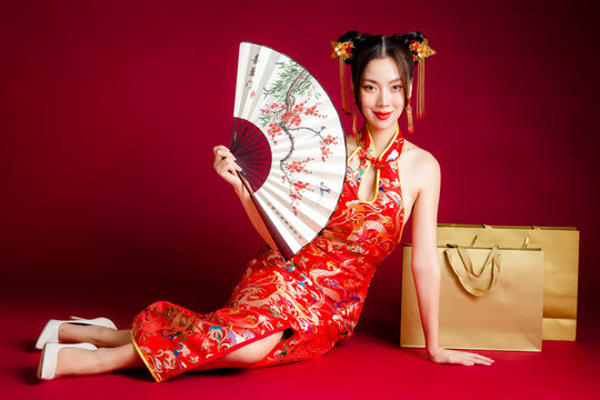 Beautiful Asian woman with clean fresh skin wearing cheongsam dress holding fan sitting with gold shopping bags on red background. Chinese text means looking for spring and happy to see plum.