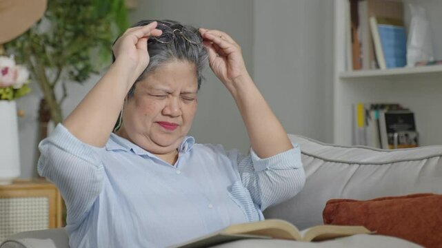 Senior woman reading book and touching glasses to stare read text,having problems with vision,sitting on sofa at home interior,door eyesight farsightedness,eye disease of elderly,health care concept