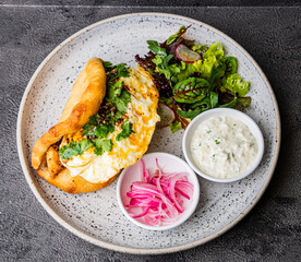 Crispy flatbread with sunny eggs, garnished with fresh cilantro, accompanied by pickled onions, creamy yogurt sauce, and a side of vibrant greens on a speckled plate.