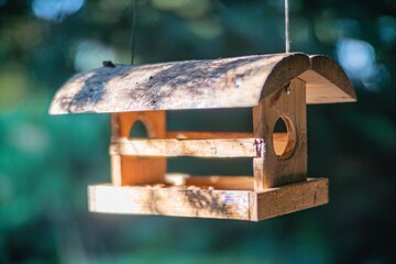 A rustic wooden bird feeder hanging from a wire, with small openings for birds, set against a blurred natural backdrop of greenery and trees