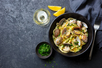 Spaghetti alle vongole, seafood pasta with clams in a black bowl and glass of wine. Dark background. Copy space. Top view.