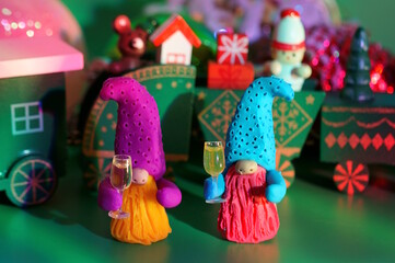 Two toy dwarfs made of plasticine with glasses of champagne on the background of a Christmas train. New Year's holidays and Christmas. Decorations and decorations. Green background.
