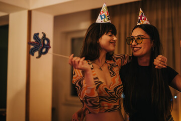 Two young women wear festive hats, smiling as they enjoy a cozy birthday celebration at home. The...