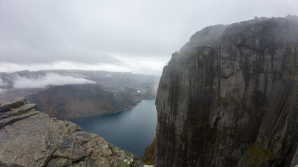 A foggy landscape featuring a steep cliff overlooking a serene fjord, surrounded by rugged mountains and misty clouds.