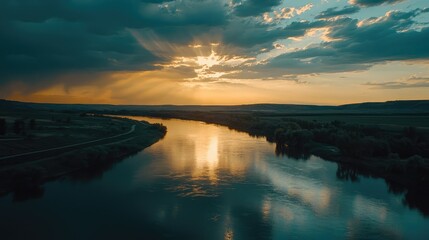 Serene Sunset Over Calm River Surrounded by Lush Greenery and Open Fields with Dramatic Cloud Patterns Illuminated by Golden Rays of Sunlight