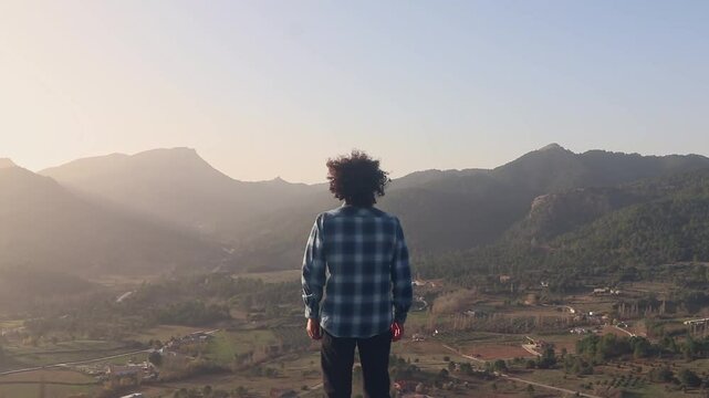 Wide shot of a man from behind with a shirt and afro hair. Man at a viewpoint reflecting on the sunset