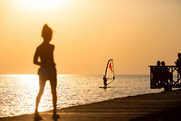 Silhouette of a woman standing on a pier at sunset, watching a windsurfer gliding across the sea