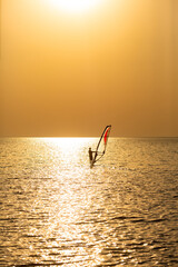 A lone windsurfer sails under the bright setting sun, creating a sparkling path on the water