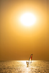 A windsurfer glides across the calm ocean under the bright sun, creating a dramatic golden glow on the water