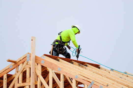 A construction worker atop a two-story wooden home under construction in a new housing division uses a power tool to install plywood on the roof structure