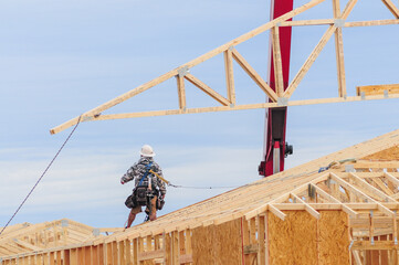 A crane lowers a roof truss over a wooden home under construction as a framing carpenter waits to guide it into place for installation