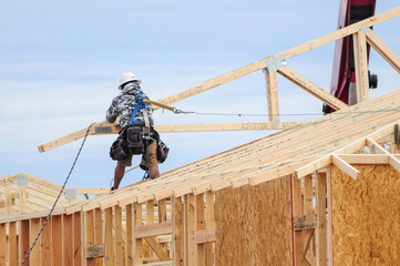 A framing carpenter atop the structure of a wooden home under construction maneuvers a roof truss...