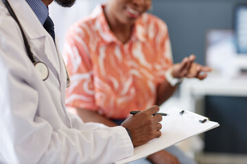 Side view close up of unrecognizable African American doctor writing notes on clipboard with woman...