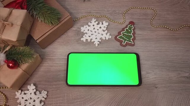 Green screen smartphone lying on wooden table horizontally next to gifts fnd DIY Christmas decorations. Christmas mockup top view.