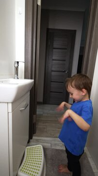 Joyful baby boy stands in the bathroom waving hands happily. Kid steps on the prop and stomps his feet actively. Child plays with a water faucet. Vertical video.