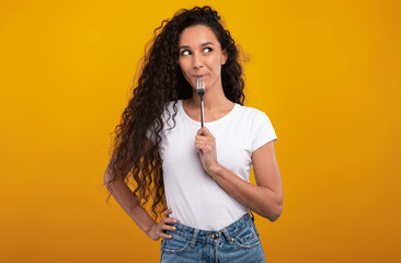 Portrait of excited hungry young woman holding fork in her mouth, biting and dreaming about...