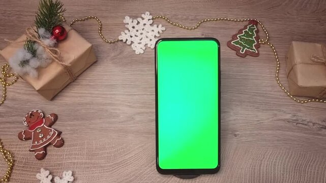 Green screen smartphone lying on wooden table next to beads , snowflakes, a Christmas tree , gifts fnd DIY Christmas decorations. Christmas mockup top view.