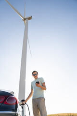 Man with charging electric car. Wind turbines in the background